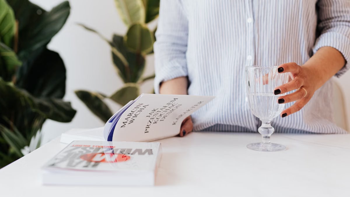 Unrecognizable female in shirt with stripes holding glass of water in hand and reading book while standing near white table